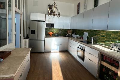 Salle lumineuse avec parquet, canapés sombres, plantes, décorations murales et grandes fenêtres laissant entrer la lumière.