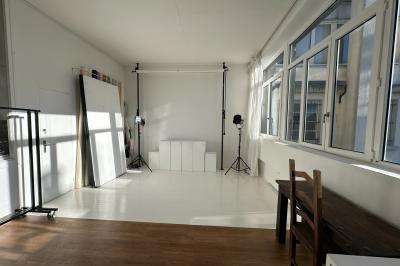 Salle lumineuse avec parquet, canapés sombres, plantes, décorations murales et grandes fenêtres laissant entrer la lumière.