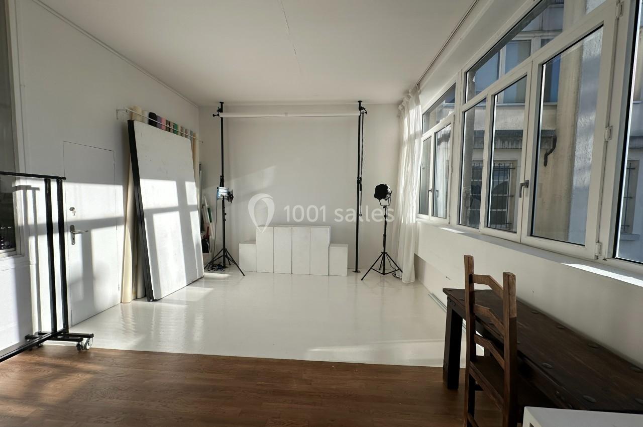 Studio lumineux avec murs blancs, grandes fenêtres, matériel de photographie et mobilier en bois minimaliste.
