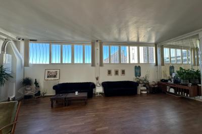 Salle lumineuse avec parquet, canapés sombres, plantes, décorations murales et grandes fenêtres laissant entrer la lumière.