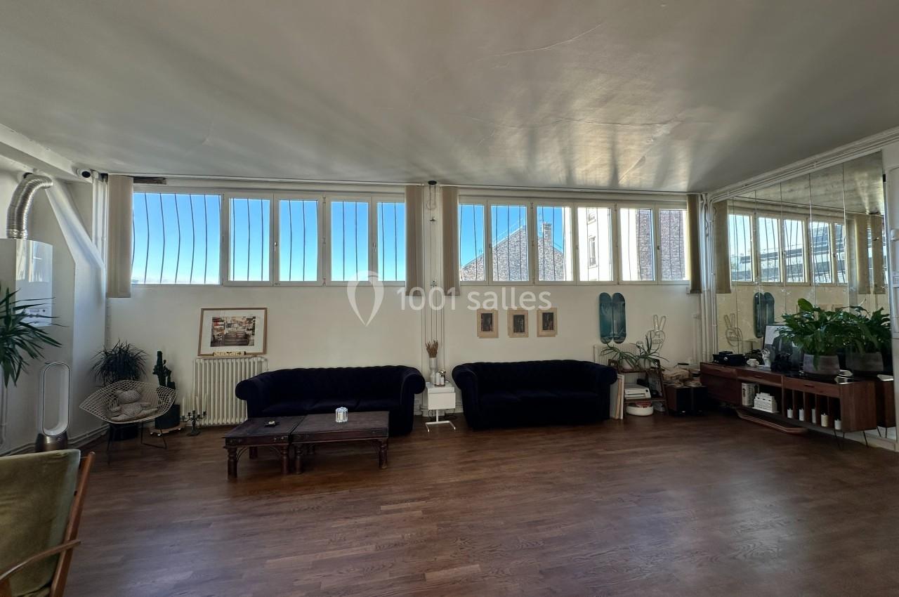 Salon lumineux avec parquet, canapés sombres, plantes, grandes fenêtres et décoration murale minimaliste.