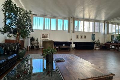 Salle lumineuse avec parquet, canapés sombres, plantes, décorations murales et grandes fenêtres laissant entrer la lumière.