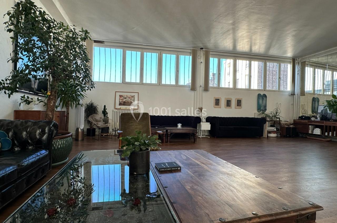 Espace intérieur lumineux avec parquet, grandes fenêtres, canapés, plantes et table basse en bois.