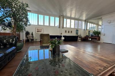 Salle lumineuse avec parquet, canapés sombres, plantes, décorations murales et grandes fenêtres laissant entrer la lumière.