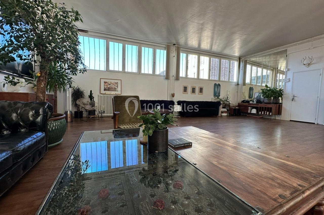 Salle lumineuse avec de grandes fenêtres, meubles en bois, canapés sombres et plantes décoratives.