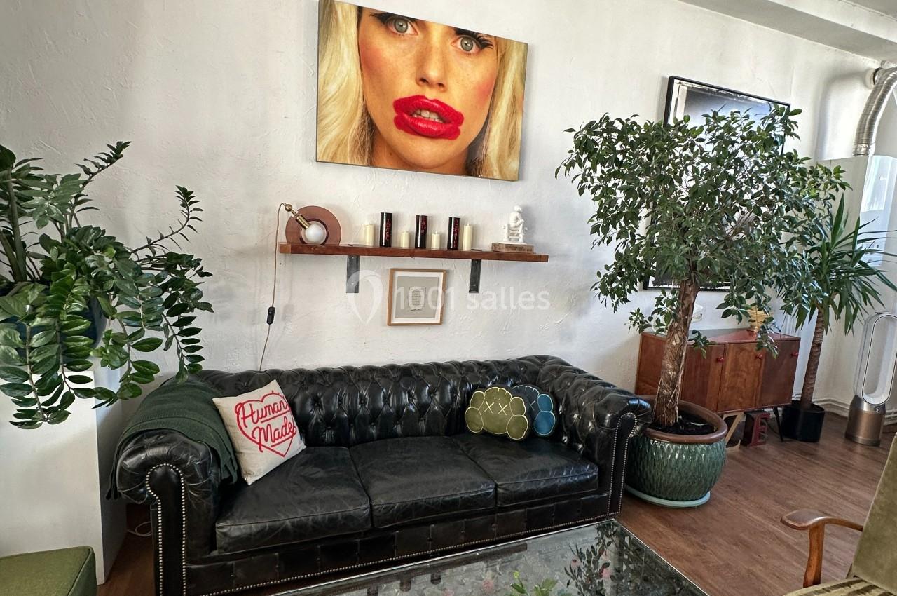 Salon avec canapé en cuir noir, plantes en pot, étagères murales et tableau représentant un visage féminin.