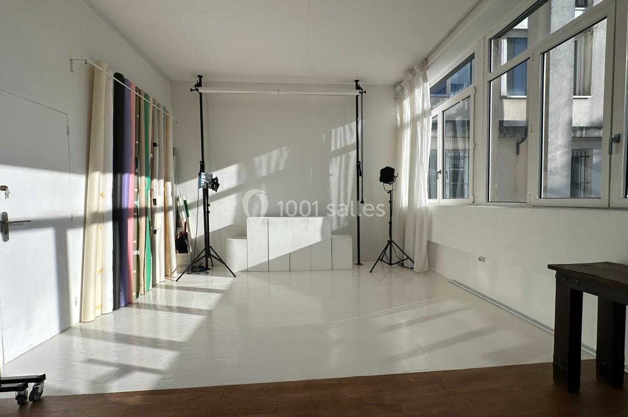 Studio lumineux avec fond blanc, trépieds pour éclairage, rideaux colorés et grandes fenêtres laissant entrer la lumière…