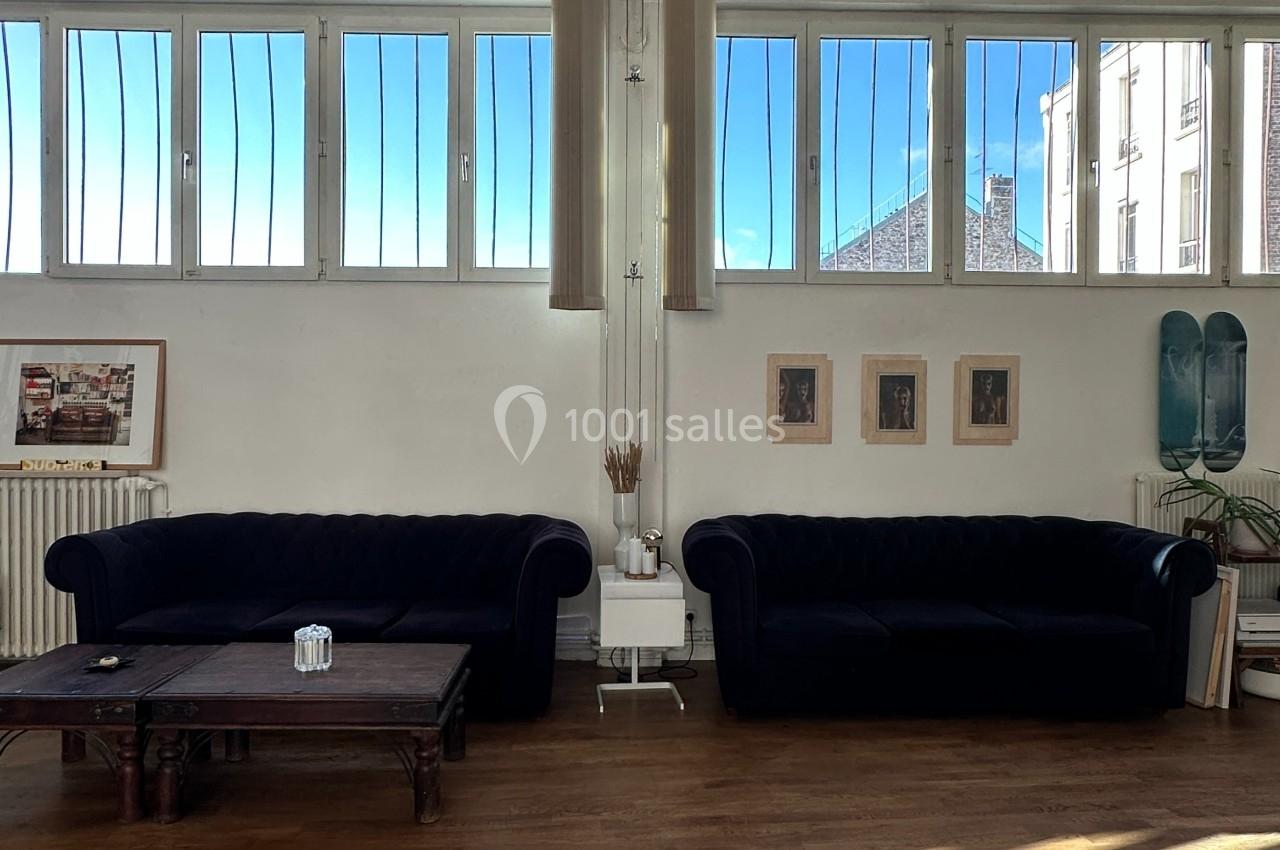 Deux canapés noirs dans un salon lumineux avec de grandes fenêtres, une table basse en bois et des décorations murales.