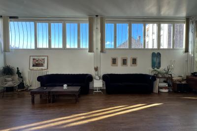 Salle lumineuse avec parquet, canapés sombres, plantes, décorations murales et grandes fenêtres laissant entrer la lumière.