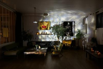 Salle lumineuse avec parquet, canapés sombres, plantes, décorations murales et grandes fenêtres laissant entrer la lumière.