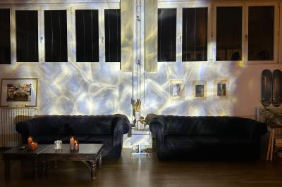 Salle lumineuse avec parquet, canapés sombres, plantes, décorations murales et grandes fenêtres laissant entrer la lumière.