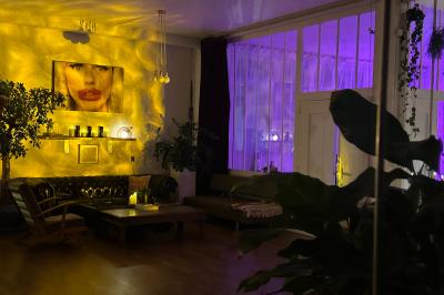 Salle lumineuse avec parquet, canapés sombres, plantes, décorations murales et grandes fenêtres laissant entrer la lumière.