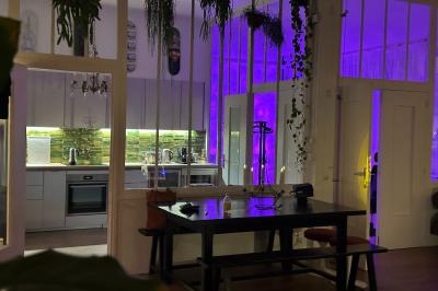 Salle lumineuse avec parquet, canapés sombres, plantes, décorations murales et grandes fenêtres laissant entrer la lumière.