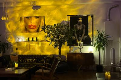 Salle lumineuse avec parquet, canapés sombres, plantes, décorations murales et grandes fenêtres laissant entrer la lumière.