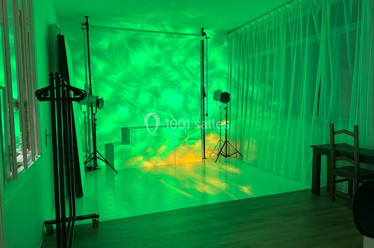 Studio photo avec éclairage vert, fond blanc, trépieds pour projecteurs et rideaux translucides sur les fenêtres.
