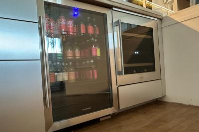 Salle lumineuse avec parquet, canapés sombres, plantes, décorations murales et grandes fenêtres laissant entrer la lumière.