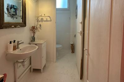 Salle lumineuse avec parquet, canapés sombres, plantes, décorations murales et grandes fenêtres laissant entrer la lumière.