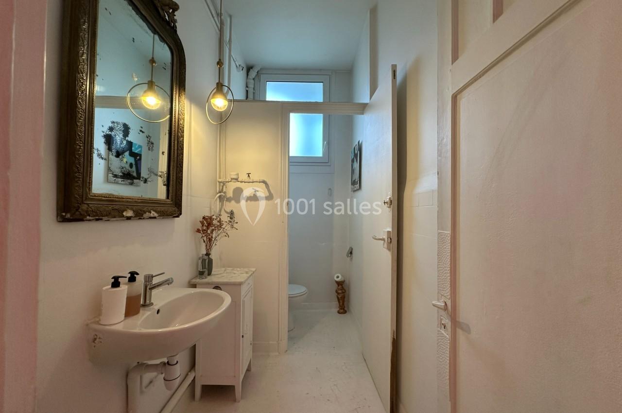 Salle de bain étroite avec lavabo, miroir ancien, toilettes au fond et lumière naturelle provenant d'une fenêtre.