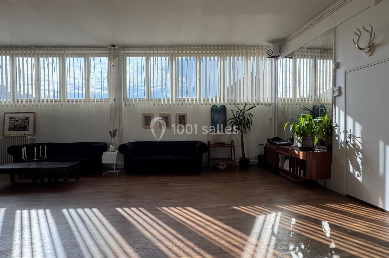 Salle lumineuse avec parquet, canapés sombres, plantes, décorations murales et grandes fenêtres laissant entrer la lumière.