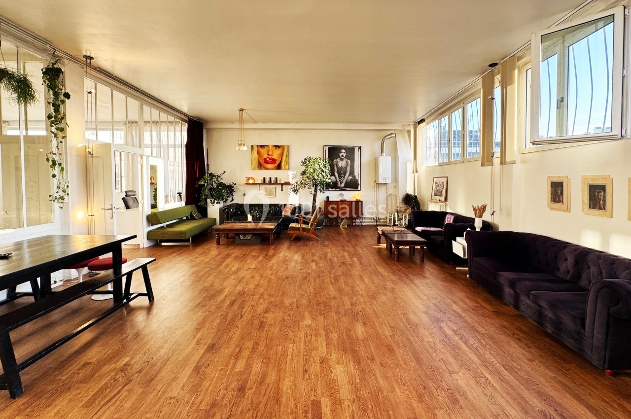 Salle spacieuse avec parquet, mobilier éclectique, plantes et grandes fenêtres laissant entrer la lumière naturelle.