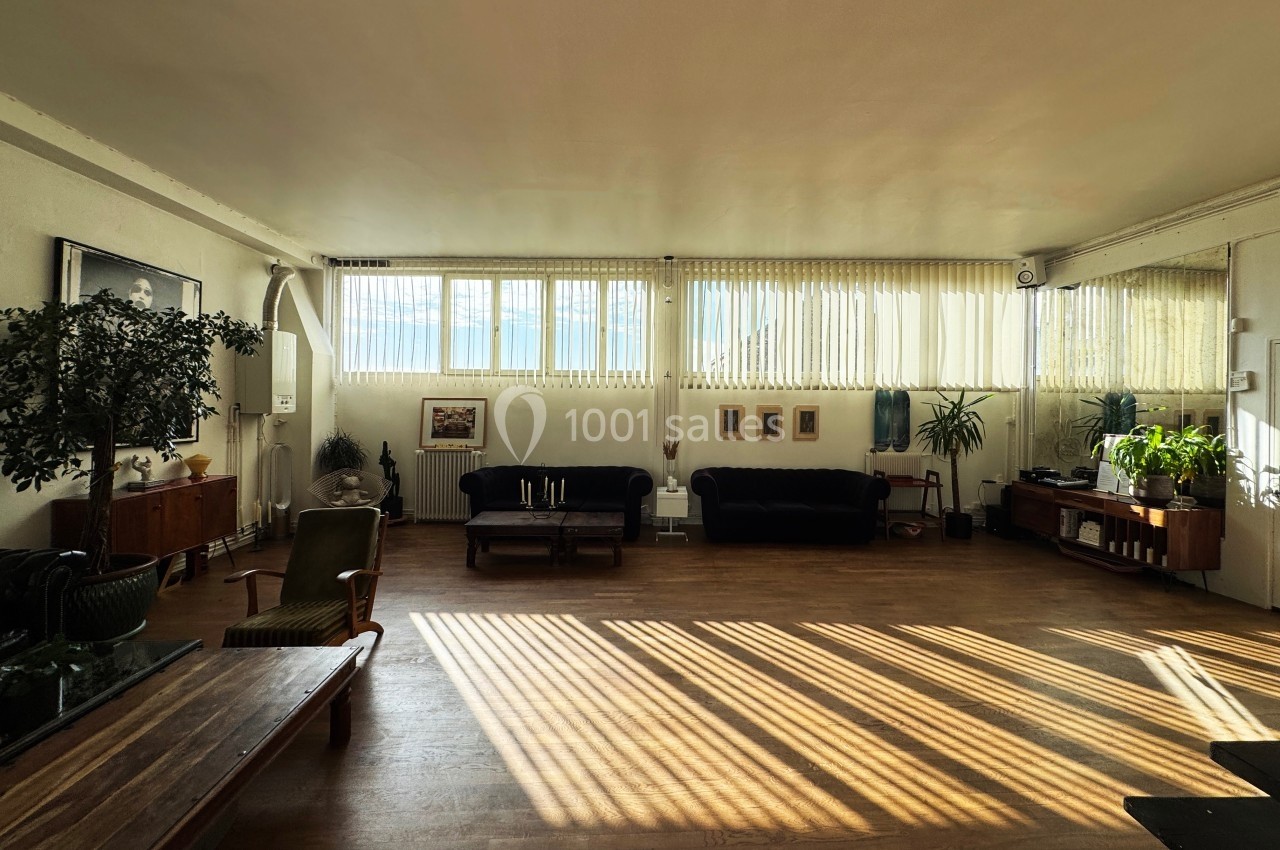 Salon spacieux avec parquet, grandes fenêtres laissant entrer la lumière, meubles sombres et plantes décoratives.