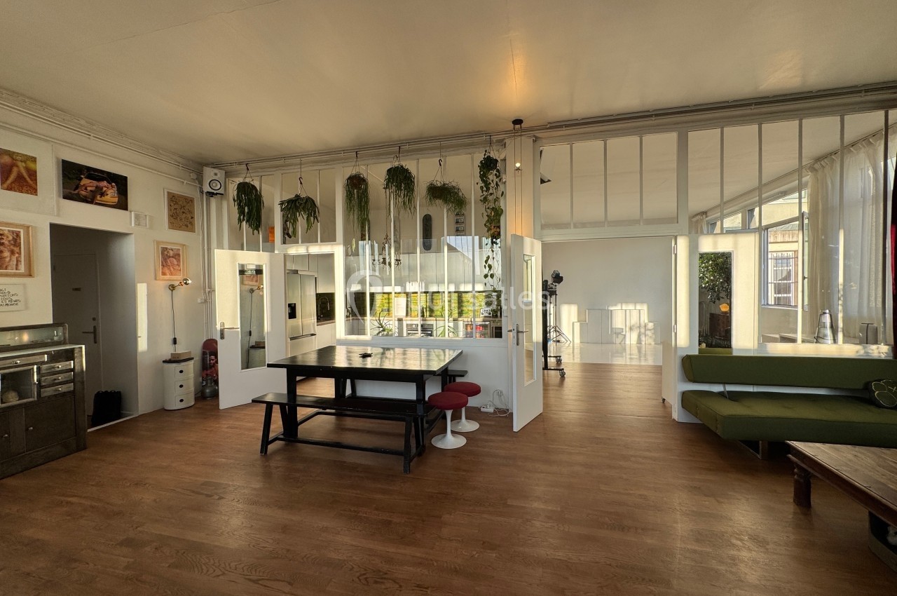 Espace intérieur lumineux avec parquet, table en bois, plantes suspendues et grandes baies vitrées.