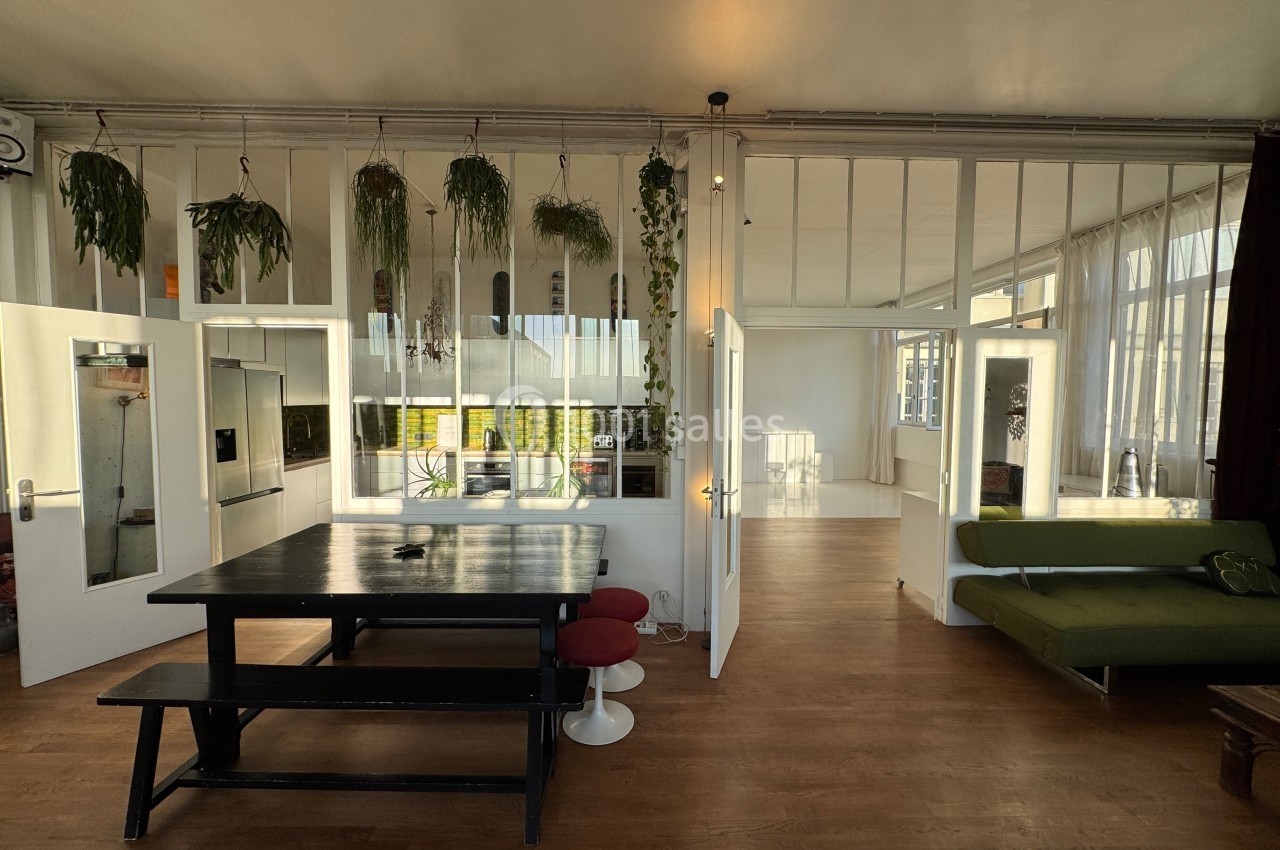 Intérieur lumineux avec table en bois, chaises rouges, canapé vert et plantes suspendues devant une verrière.