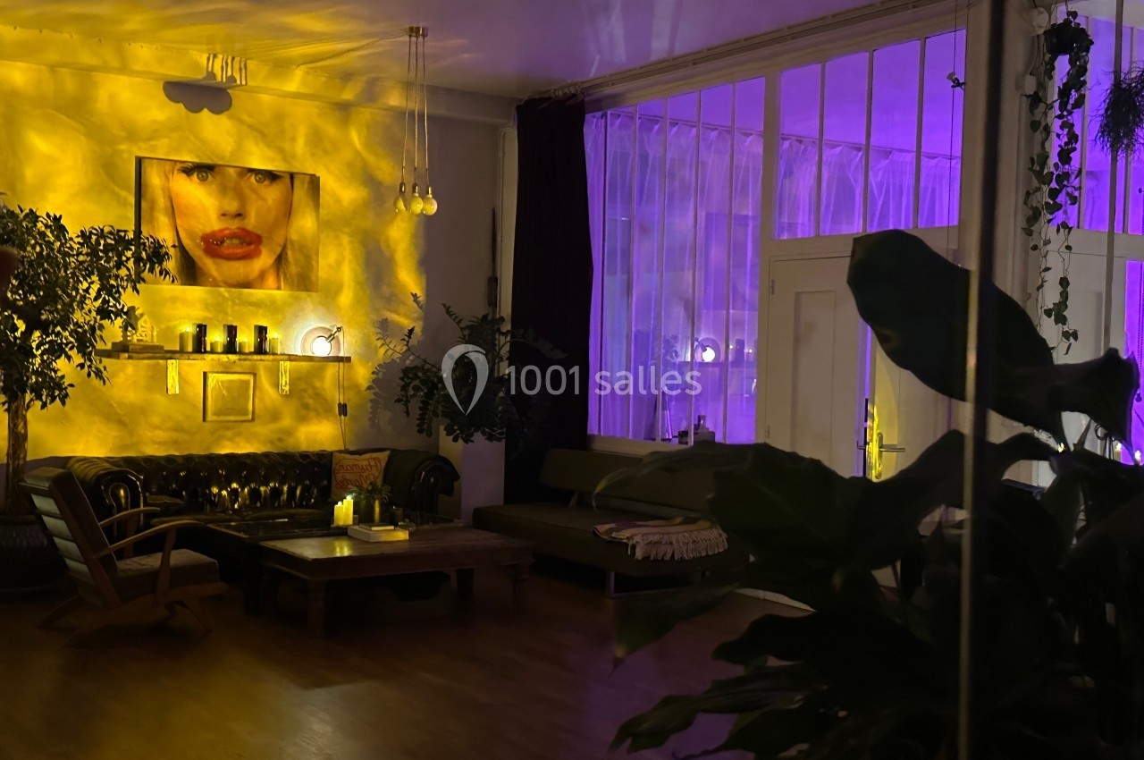 Salon cosy éclairé par une lumière tamisée violette et jaune, avec plantes, canapé en cuir et décoration murale.