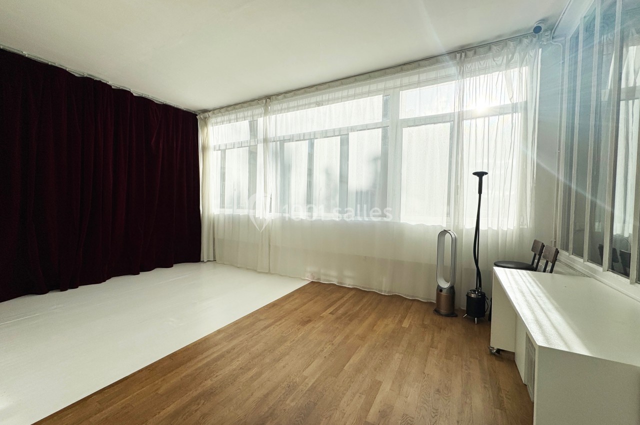 Salle lumineuse avec parquet, rideaux rouges, murs blancs et grandes fenêtres laissant entrer la lumière naturelle.