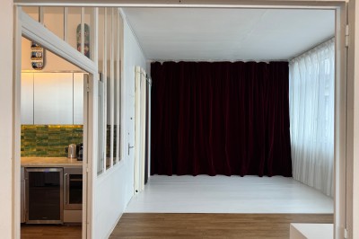 Rideaux rouges fermés éclairés par une lumière rose dans une pièce aux murs blancs avec des voilages translucides.