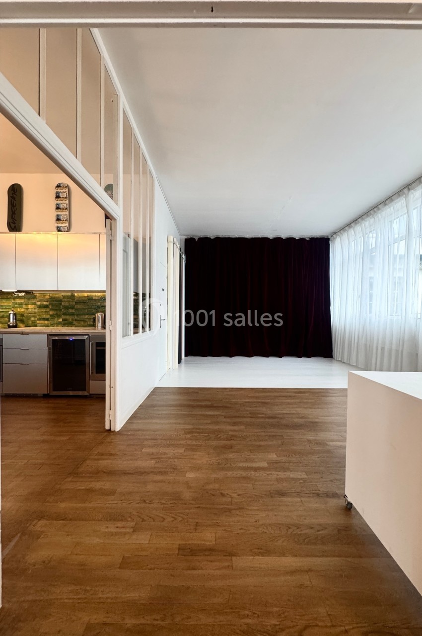 Vue d'une pièce lumineuse avec parquet, cuisine ouverte à gauche, rideaux sombres et grandes fenêtres à droite.