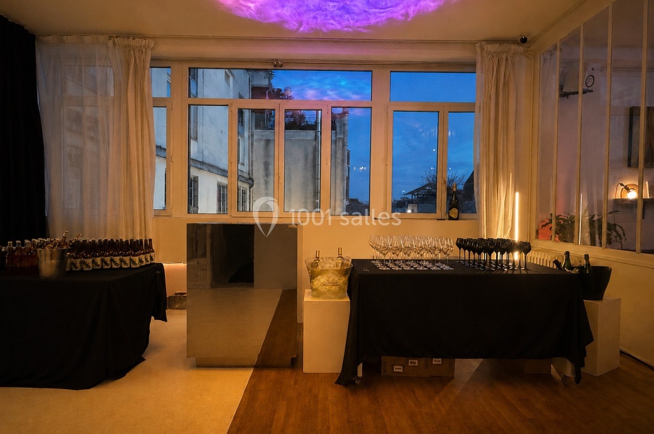 Salle de réception avec grandes fenêtres, tables dressées avec bouteilles, verres et éclairage d'ambiance tamisé.