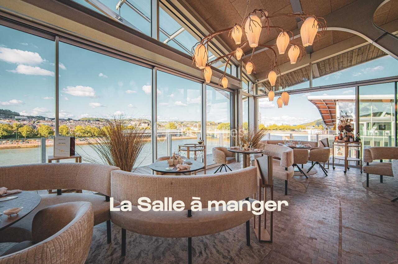 Salle à manger lumineuse avec grandes baies vitrées offrant une vue sur un fleuve et des collines verdoyantes.