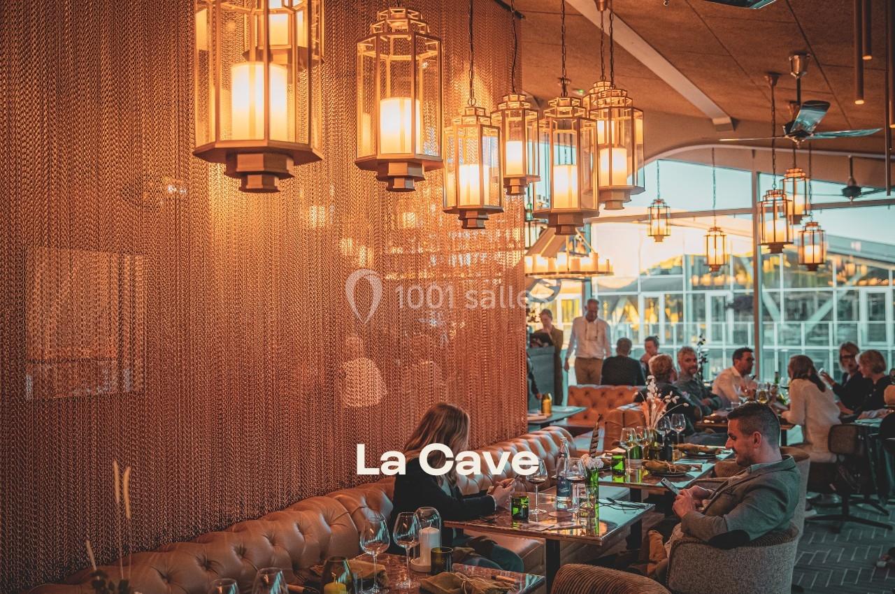 Salle de restaurant chaleureuse avec éclairage tamisé, tables occupées et grandes baies vitrées en arrière-plan.