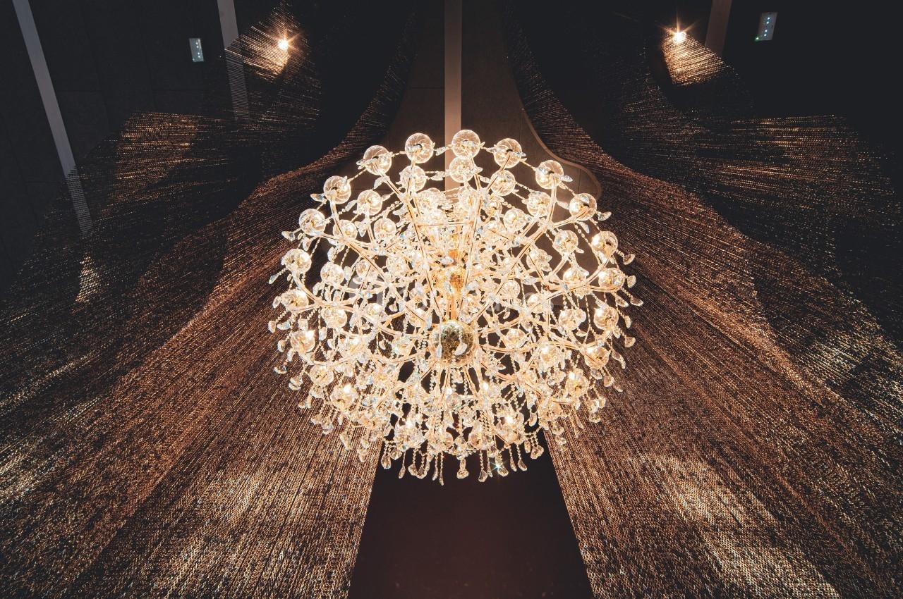 Lustre en cristal suspendu, éclairé, avec un fond sombre et des rideaux texturés.