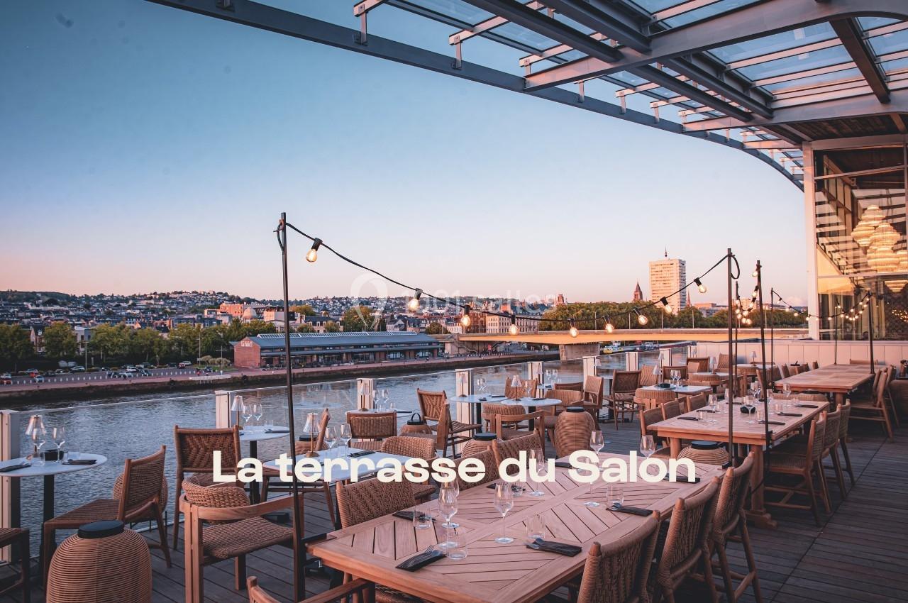 Terrasse en bois avec tables dressées, vue sur un fleuve et une ville au coucher du soleil.