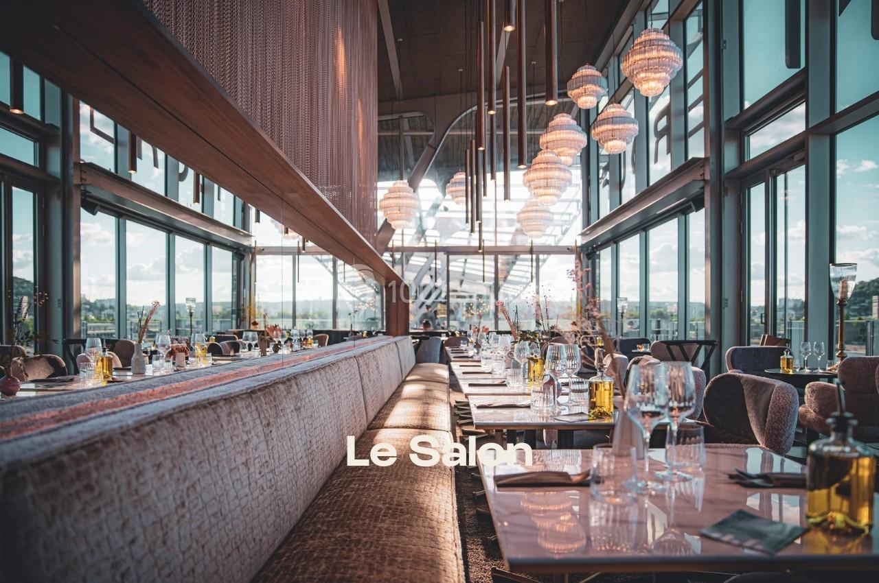 Salle de restaurant lumineuse avec de grandes baies vitrées, tables dressées et suspensions décoratives.