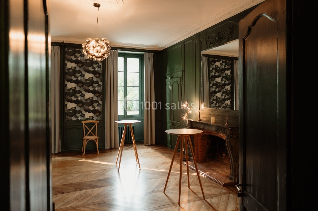 Salle élégante avec parquet en bois, murs verts, cheminée, fenêtres avec rideaux à motifs et tables hautes en bois.