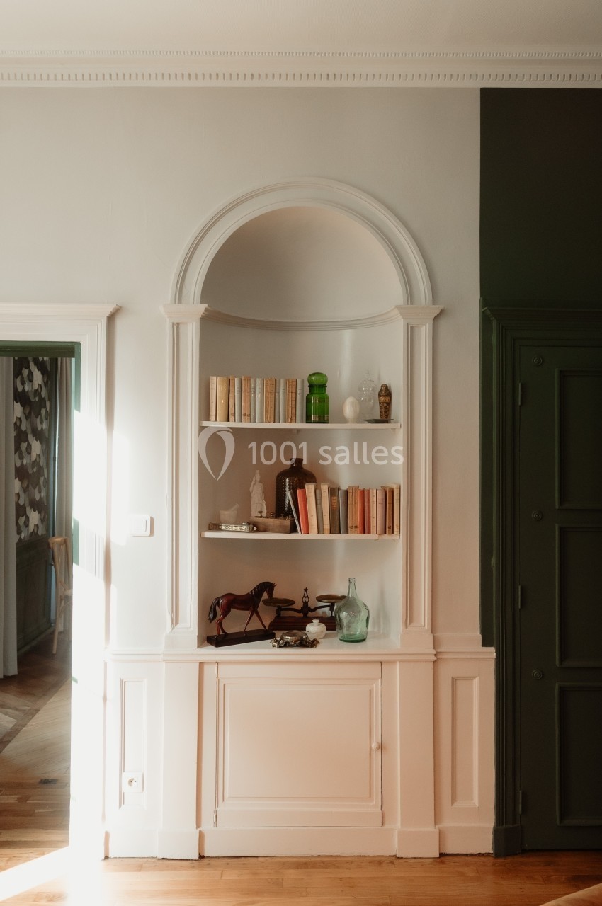 Niche murale avec étagères contenant des livres, objets décoratifs et lumière naturelle entrant par une porte à gauche.