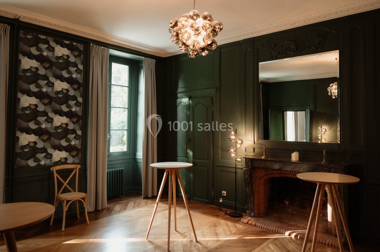 Salle élégante avec murs verts, parquet en bois, cheminée, miroir, tables hautes et lumière naturelle entrant par la fenêtre.