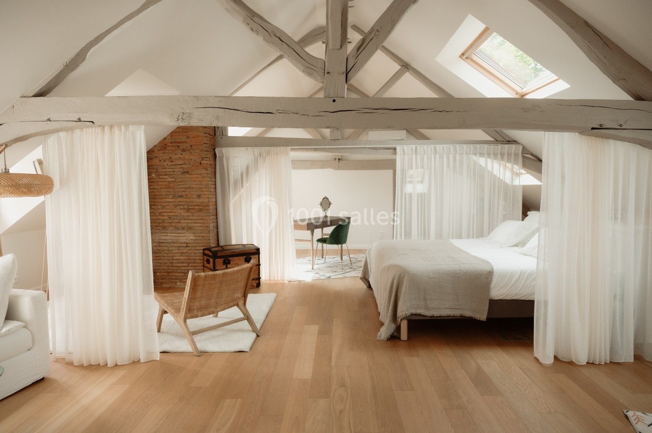 Chambre mansardée lumineuse avec poutres apparentes, lit double, bureau, fauteuil en bois et rideaux blancs.