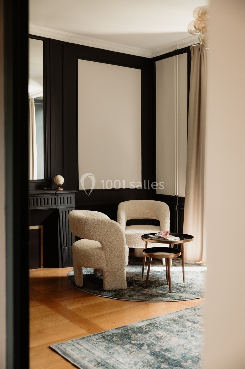 Deux fauteuils en tissu clair autour d'une table basse sur un tapis bleu, dans un salon aux murs sombres et rideaux beiges.