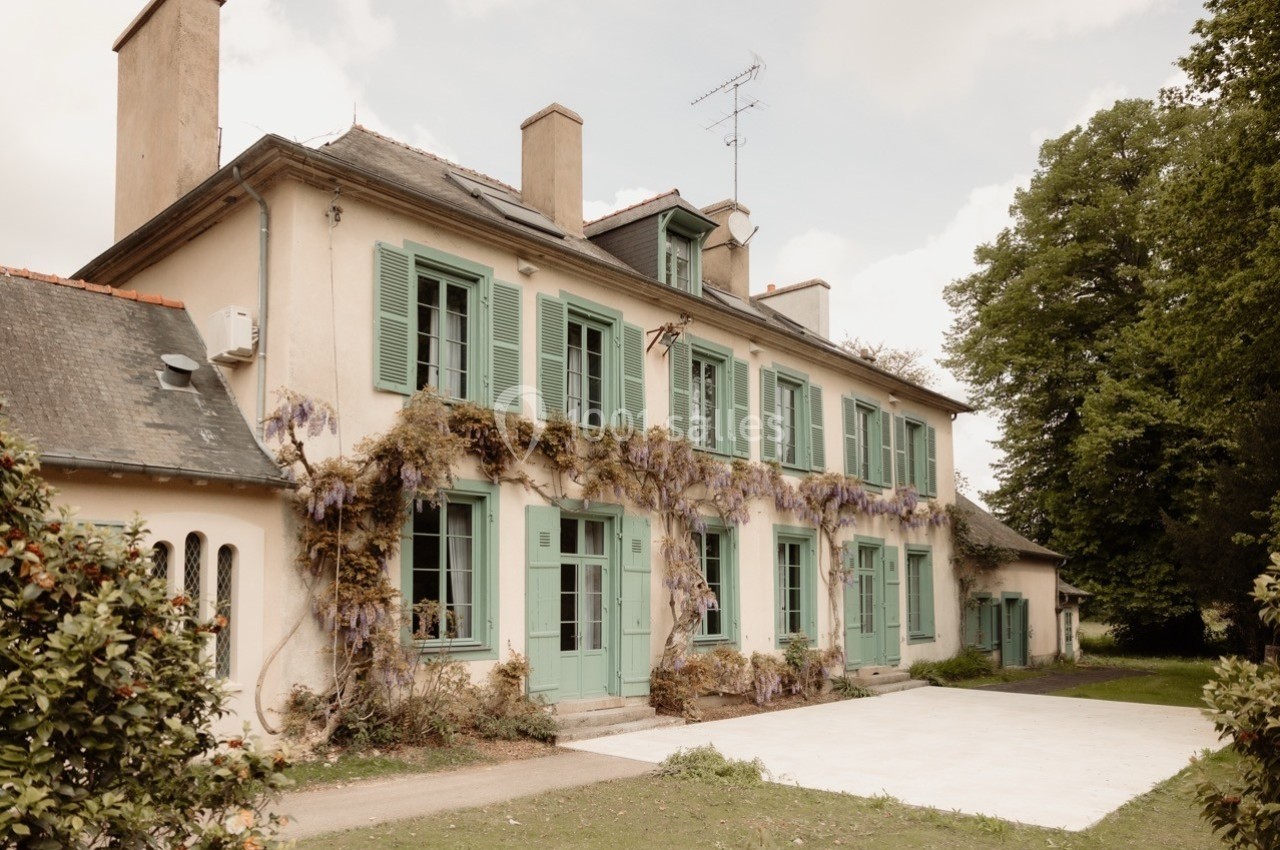 Façade d'une maison ancienne avec volets verts, ornée de glycines, entourée de verdure et d'un ciel partiellement nuageux.