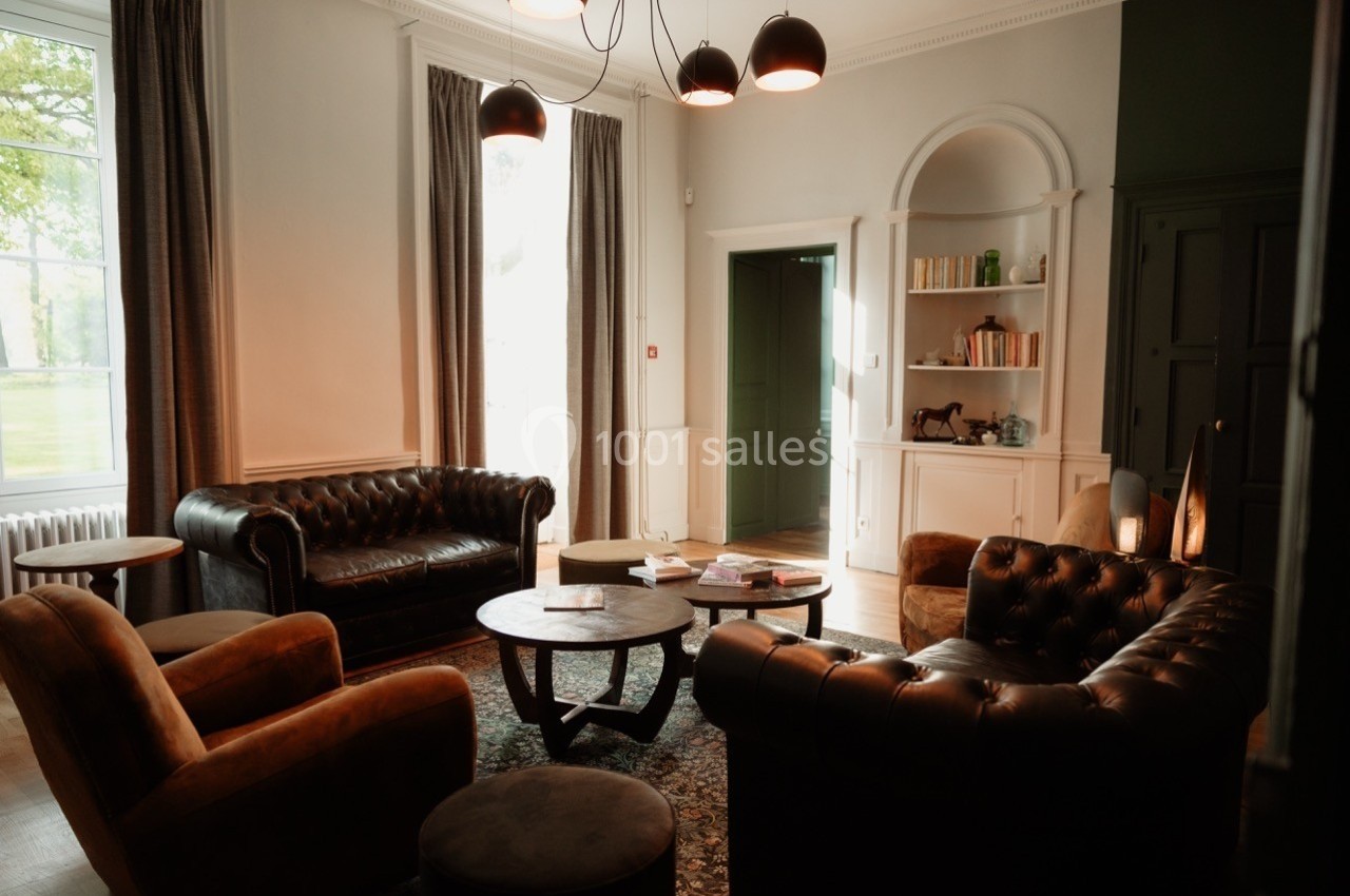 Salon lumineux avec canapés en cuir, fauteuils, tables basses, étagères et grandes fenêtres donnant sur un jardin.