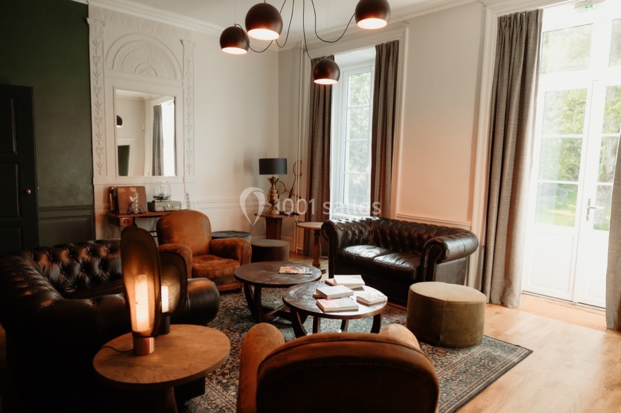 Salon lumineux avec fauteuils en cuir, tables basses, lampes modernes et grandes fenêtres donnant sur un jardin.