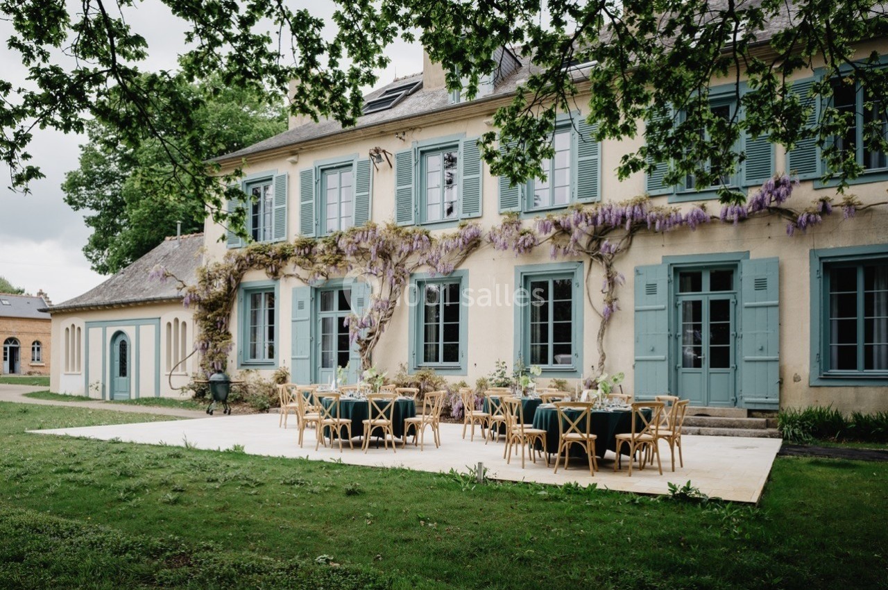 Façade d'une maison ancienne avec volets bleus, terrasse aménagée et glycines grimpant sur les murs.