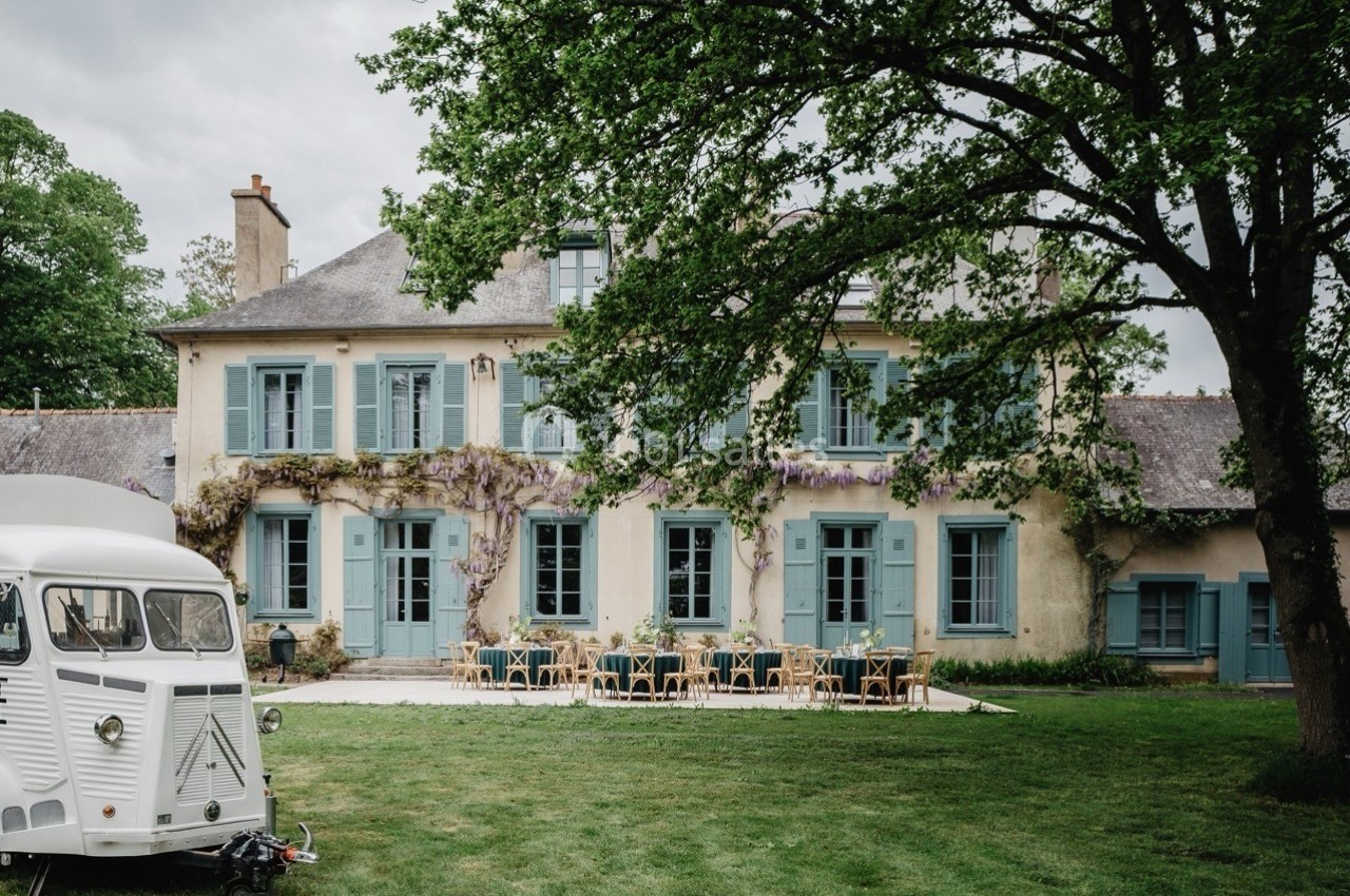 Façade d'une maison ancienne avec volets bleus, terrasse aménagée et un van blanc stationné sur la pelouse.
