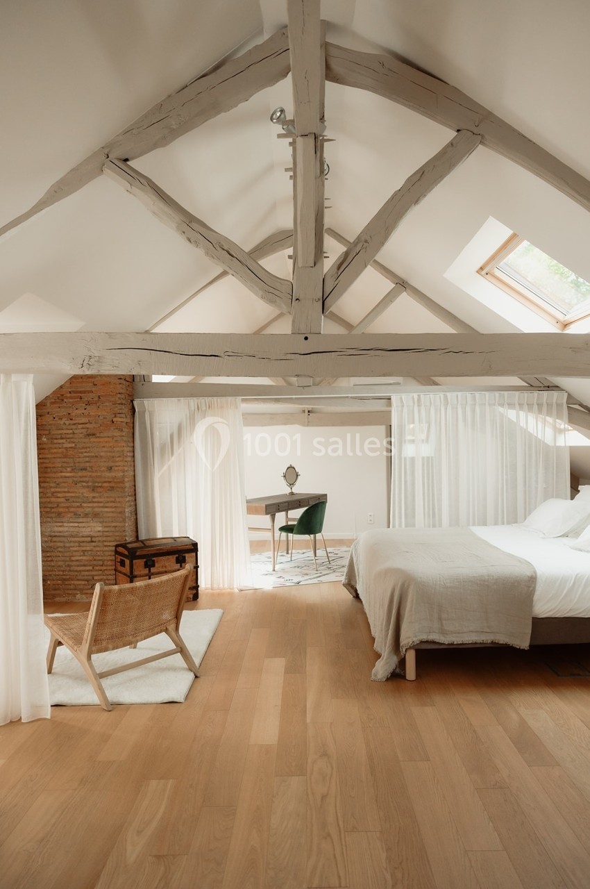 Chambre mansardée lumineuse avec poutres apparentes, lit double, chaise en bois, bureau et coffre ancien.