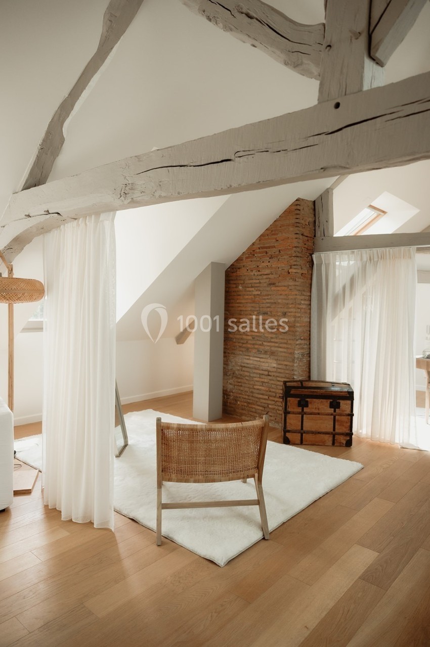 Espace lumineux sous combles avec poutres apparentes, chaise en rotin, tapis blanc et mur en briques.