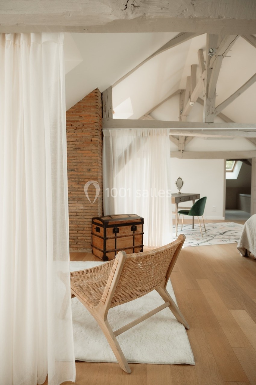 Fauteuil en rotin sur un tapis clair dans une chambre mansardée avec poutres apparentes, coffre en bois et bureau.